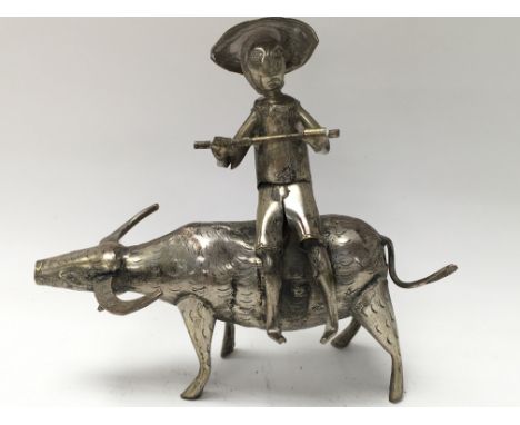 A white metal figure of an oriental man holding a stick whilst sat on a water buffalo.