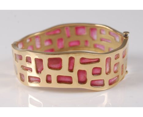 A yellow metal hinged oval bangle, featuring a wavy design with assorted apertures filled with red glass inlay, bangle diamet