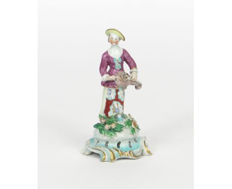 A Bow figure of a female musician, c.1760-65, standing and playing the hurdy-gurdy, raised on a tall scrolled base applied wi