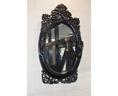 AN EARLY 20TH CENTURY EBONISED AND CARVED HARDWOOD OVAL WALL MIRROR, the heavily carved finial and base with lattice stole, f