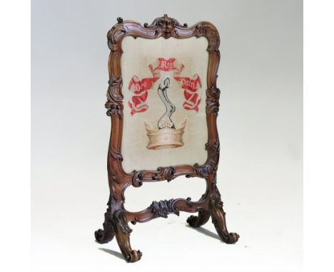A Victorian carved walnut firescreen, inset with a woolwork family crest, within a scrolled surround, surmounted by a mask, o