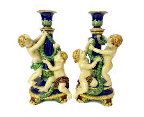 PAIR OF MINTON MAJOLICA FIGURAL CANDLESTICKS, each surmounted with a pair of putti playing with a garland, on oval bases with