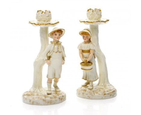 A PAIR OF ROYAL WORCESTER GLAZED PARIAN WARE BOY AND GIRL CANDLESTICKS MODELLED BY JAMES HADLEY IN THE STYLE OF KATE GREENAWA