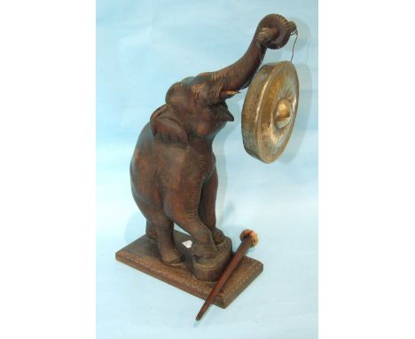 An early-20th century carved wood elephant with raised trunk, holding a brass dinner gong, 51cm high, (one tusk damaged).