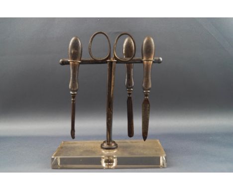 A silver mounted steel four piece manicure set with engine turned handles, on a silver stand with a clear perspex rectangular