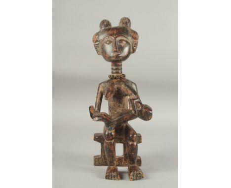 A CARVED AFRICAN FIGURE OF A WOMAN SITTING ON A BENCH, feeding a baby. 14ins high.