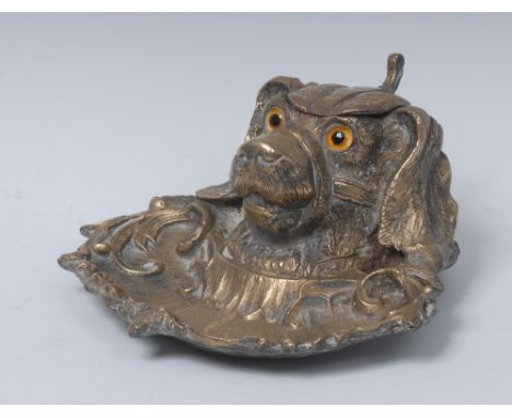 A 19th century brass novelty inkwell, cast as the head of a dog, hinged cover, glass eyes, she'll shaped base cast with leafy