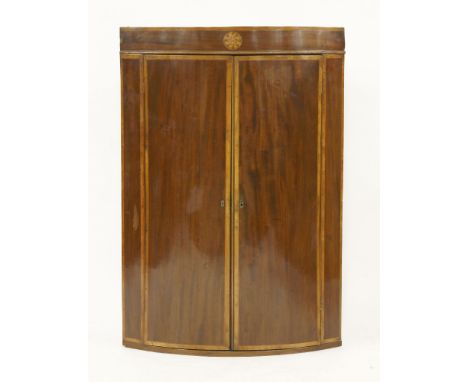 A George III crossbanded mahogany bow fronted hanging corner cabinet, the plain cornice with an inlaid sunburst, 76cm wide, 1