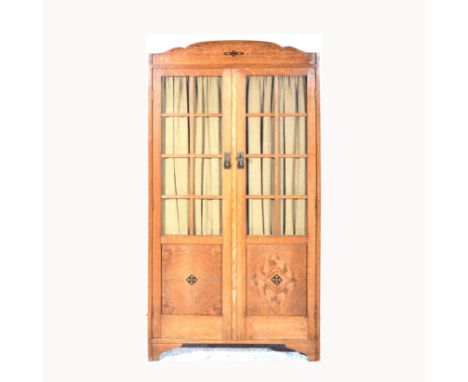 An Arts & Crafts oak bookcase cabinet, arched top, glazed and panelled doors, inlaid with stylised motifs, enclosing four adj