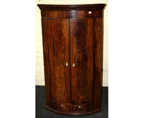 A late George III figural mahogany convex hanging corner cupboard enclosed by a pair of panel doors having two drawers below.