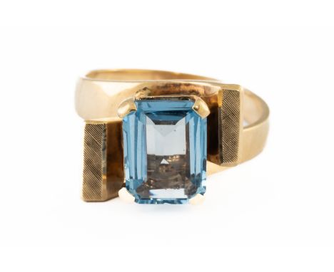 A GOLD AND BLUE TOPAZ SINGLE STONE RINGMounted with a rectangular step cut blue topaz between textured twistover shoulders, d