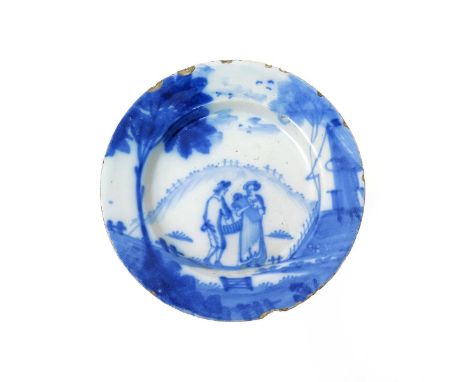 A delftware plate  mid 18th century, painted in blue with a couple standing before a hillside beneath a tall tree, she cradli