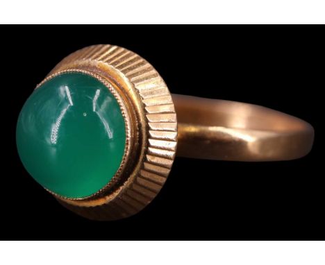 A 1960s jade finger ring, having an 8 mm cabochon, set on a radially sawtooth-stepped bezel, protuberant to a yellow metal ba