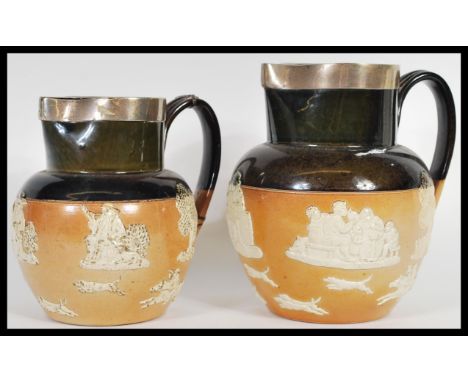 A graduating pair of Royal Doulton Lambeth harvest ware salt glazed jugs having silver hallmarked marked rims. Decorated with