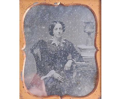 A Victorian leather cased daguerreotype, depicting a three-quarter length portrait of a woman seated at a table beside an urn