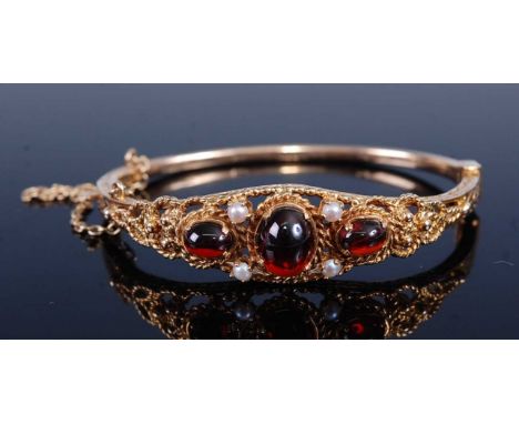 A 9ct yellow gold, garnet and pearl hinged bangle, featuring a rope edge openwork panel with three garnet cabochons and four 