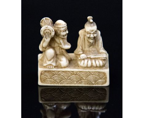 An Oriental ivory netsuke, carved as two figures seated on a plinth, one with a drum, the other with a fan, width 3.5cm