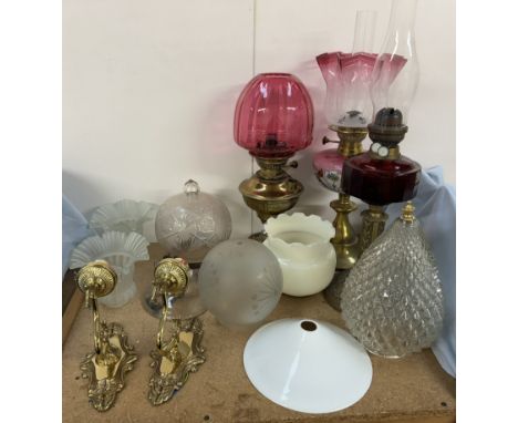 A cranberry glass oil lamp on a brass column together with two other oil lamps, a pair of wall lights and glass light fitting