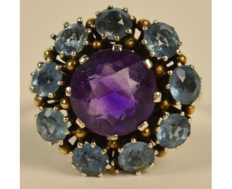 A white metal floral set ring, the central amethyst coloured stone in a border of pale blue stones in pierced scrolling mount