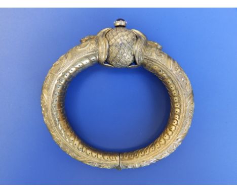An Eastern 19thC gilt metal bangle, having raised floral scroll decoration, a screw-thread pin with red enamel head allowing 