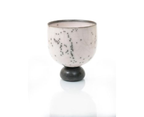 •Tim Andrews (born 1960) a raku footed cup form,  the pale pink/off white open bowl with black spot decoration, on black comp
