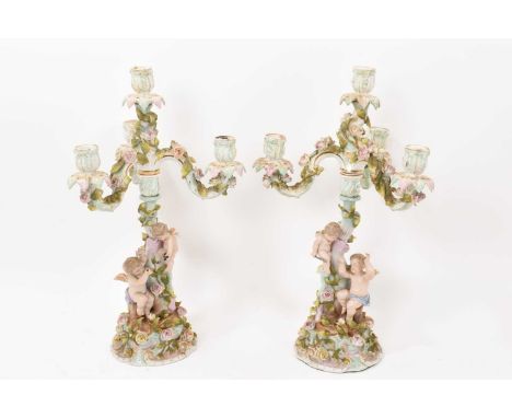 A pair of Dresden porcelain three branch candelabra, decorated with cherubs around a central tree, with floral encrusted orna