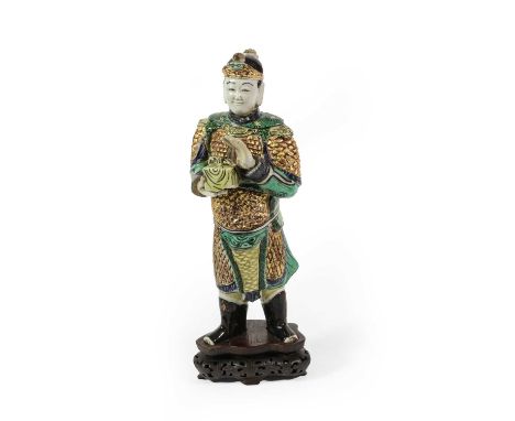 A Chinse Porcelain Figure of a Warrior, Kangxi, standing wearing armour, a box in his right hand, painted in famille rose ena