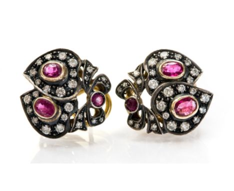 A pair of ruby and diamond earrings, each with two overlapping hearts topped with a bow, collet set oval rubies surrounded by