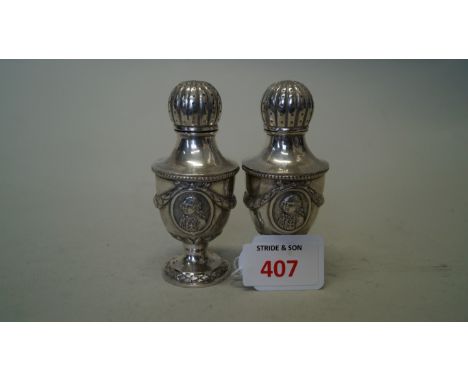 A pair of continental white metal urn shaped pepperettes, decorated portrait plaques, swags and ribbons, 10cm.