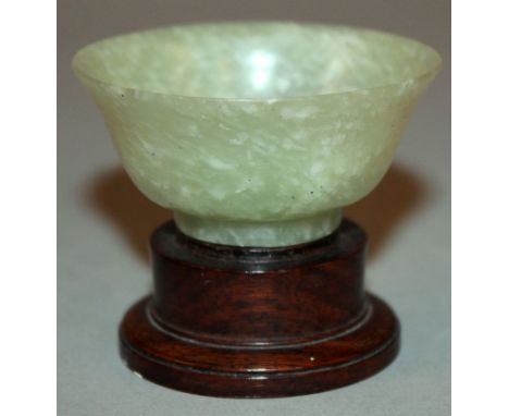 AN EARLY 20TH CENTURY CHINESE JADE-LIKE CELADON GREEN WINE CUP, with a wood stand, the stone with white inclusions, the cup i