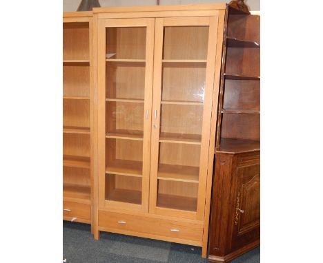 A light oak display cabinet, with two glazed doors enclosing four shelves, above a single frieze drawer, raised on square blo