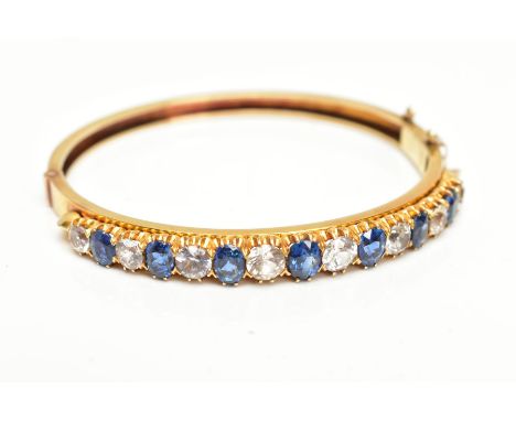 A YELLOW METAL GEMSET BANGLE, yellow metal hinged bangle set with seven oval cut blue sapphires interspaced with eight colour