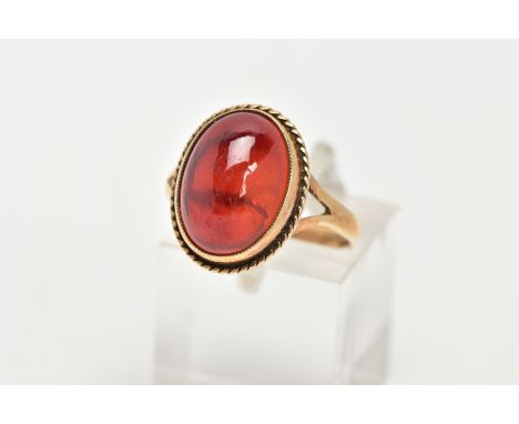 A 9CT GOLD RING, designed with a hollowed out, oval red cabochon, bezel set with a rope twist surround, bifurcated shoulders 