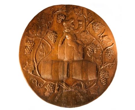 An 18th century circular oak barrel end, carved with bacchanalian putto, wearing feathered head band holding glass aloft in h
