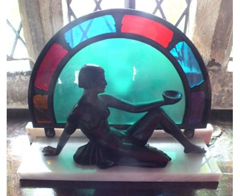 An Art Deco design table lamp, fashioned as a bronzed cast metal female seated on a white marble base, before a stained glass