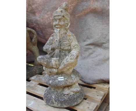 A weathered cast composition stone garden ornament in the form of a seated cross legged gnome smoking a long handled pipe wit