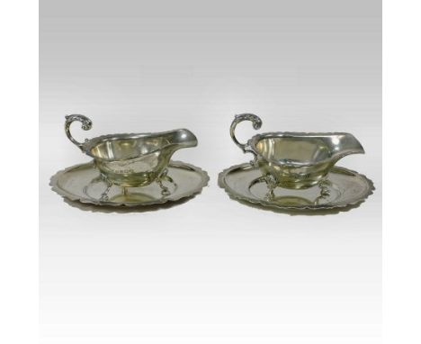 A pair of early 20th century silver sauce boats on stands, each of helmet shape, with a flying scrolled handle, on three hoof