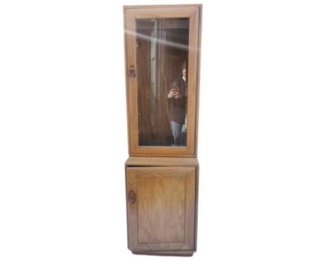 A light elm Ercol glazed tall cabinet, with single glazed door above a cupboard enclosing a single shelf, with manufacture ma