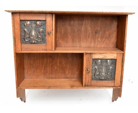 An Arts &amp; Crafts light oak hanging shelf, with two cupboard doors inset with embossed copper coloured metal panels, heigh