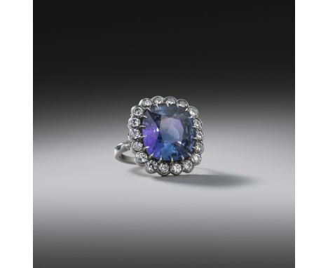 A colour-change sapphire and diamond cluster ring, the cushion-shaped sapphire weighing 10.90cts, claw-set within a surround 