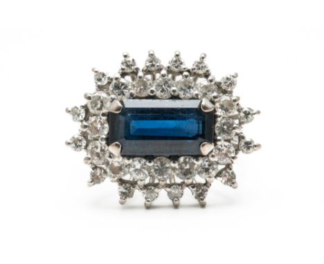 A SAPPHIRE AND DIAMOND CLUSTER RINGClaw set with the rectangular emerald cut sapphire in a two row surround of graduated roun