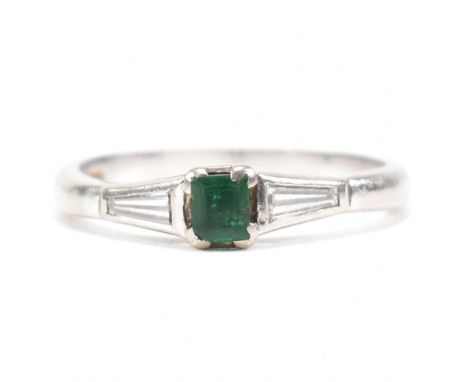 A hallmarked 18ct white gold, emerald and diamond ring. The ring having a central step cut emerald flanked by two bezel set t