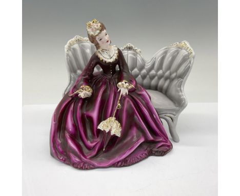 A semi-porcelain figure modeled as a brunette lady wearing a purple grown with gilded decorations sitting on couch with paras