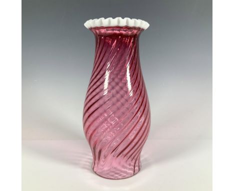 A cranberry colored glass lamp shade with a round body that narrows out, featuring a spiral design and a white ribbon pattern