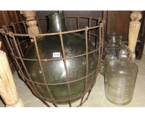 A vintage green glass carboy encased in a wirework basket together with a five litre glass measure plus further jar of squat 