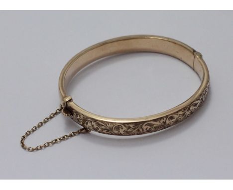 A 9ct gold stiff hinged bangle, the top half worked with a scrolling, floral design and clasp with safety chain. Gross weight