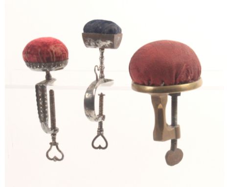 Three metal sewing clamps comprising a brass example with steel screw below a circular pin cushion top, 15cm, a steel example