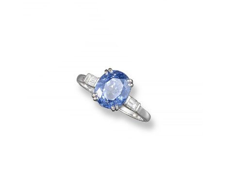 A sapphire and diamond ring, the oval-shaped sapphire is set with a baguette-shaped diamond to each shoulder in platinum. Siz