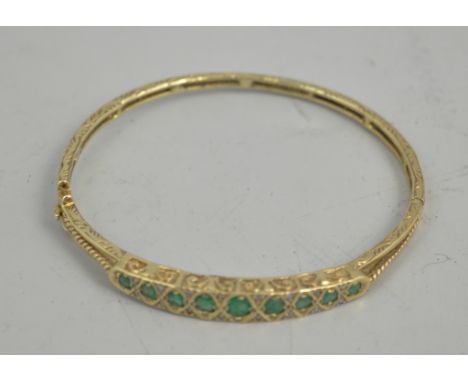 Yellow metal hinged bangle, set with nine graduated emeralds and small diamonds, engraved.