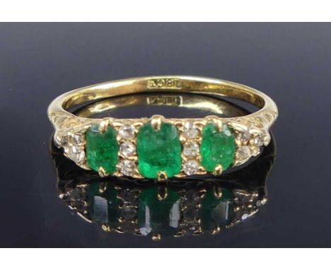 An 18ct gold, emerald and diamond ring, in carved and pierced elliptical setting, arranged as three oval cut emeralds (worn a